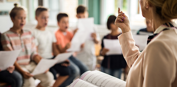 Teacher standing with music book open in front of young school-age children singing