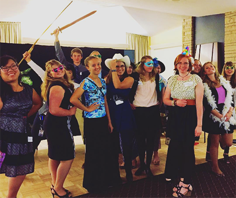 Lower Division Students holding props, some wearing masks in front of a photo wall backdrop
